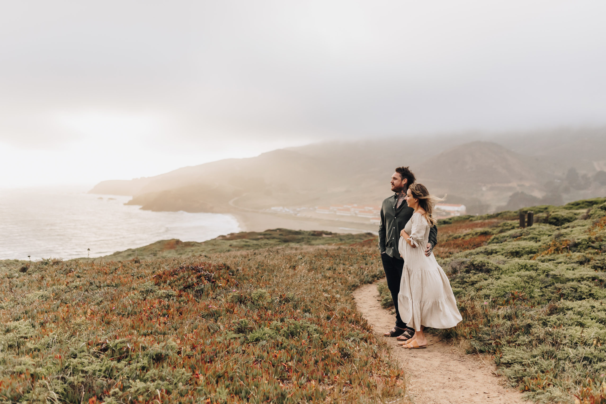 A Couple looks out over the ocean, she is pregnant and they both have tatoos