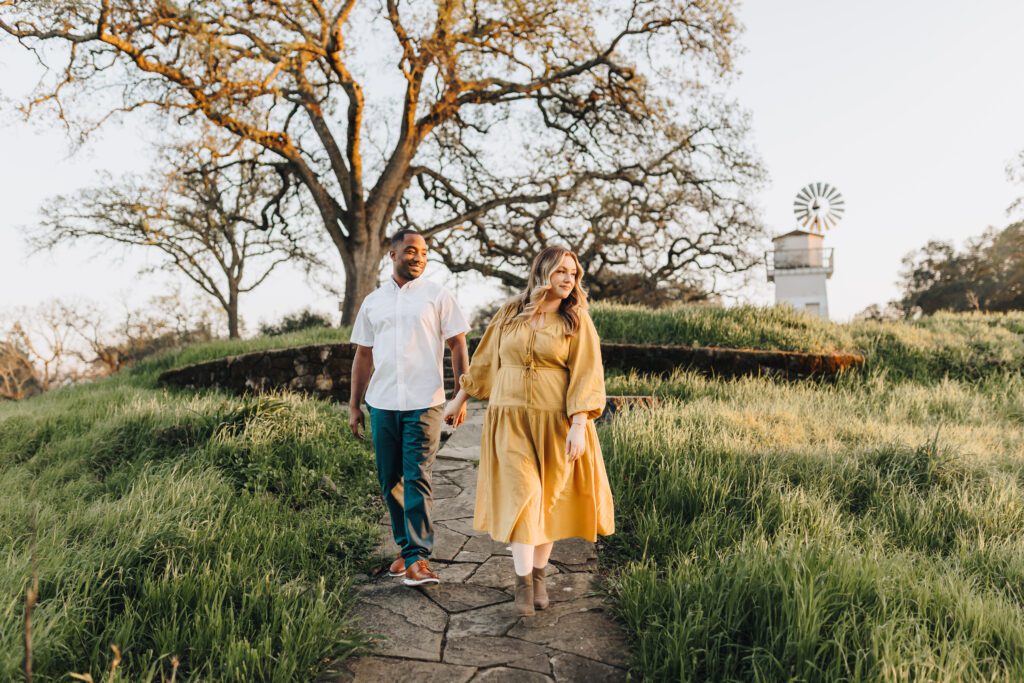 A Couple walks down a path bordered by green grass with a beautiful Oak tree and windmill behind them. They are in Walnut Creek, CA. 