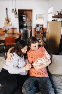 Family sits on couch with newborn baby, mom and dad are smiling at baby