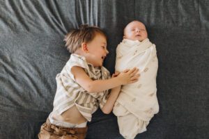 Big brother laughs while laying on the bed with swaddled baby sister