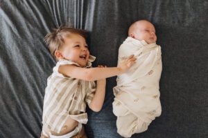Big brother laughs while laying on the bed with swaddled baby sister