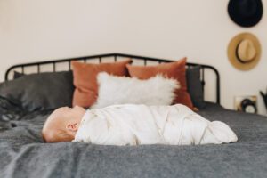 Baby sister lays on the bed swaddled and asleep during lifestyle newborn session