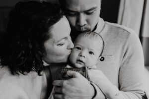 Black and white of Mom and dad kissing baby who looks at the camera