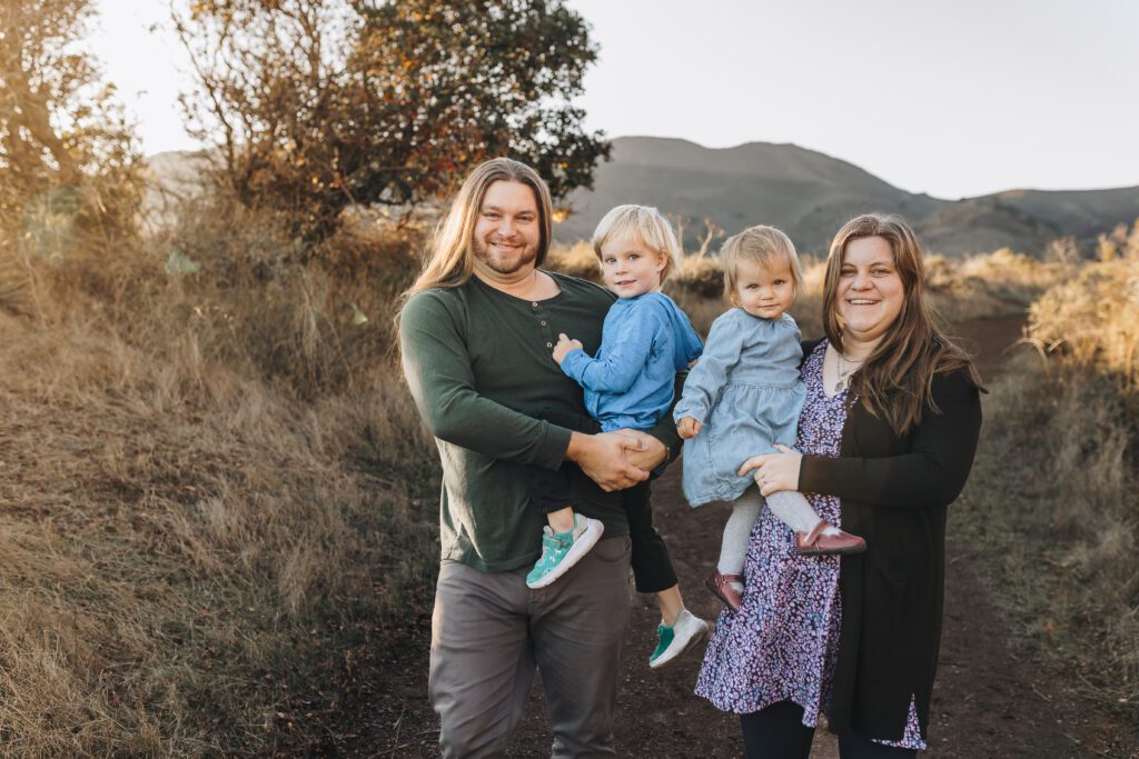 A family of four with a mom and dad and two kids smiles at the camera as the sun kisses them. 