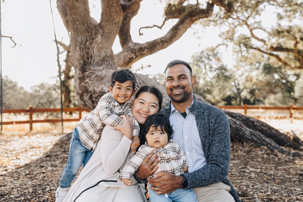 A family of four sits under a big oak tree as the sun comes up behind them, and the smile at the camera, all snuggled together in Walnut Creek. 